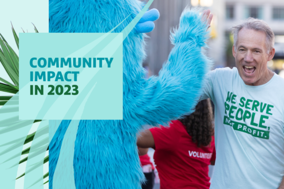 Smiling man high-fiving a friendly, blue monster mascot at the finish line of a run with text headline "Community Impact in 2023" overlaid.