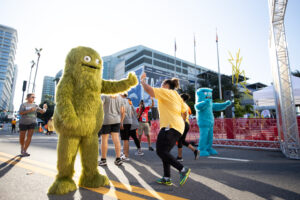 Participants in a fun run high-five colorful monster mascots on a city street, with event structures and flags in the background.