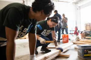 Two people wearing 'We Serve People, Not Profit' t-shirts are working together to measure and mark a wooden plank in a workshop setting with tools and equipment around them.